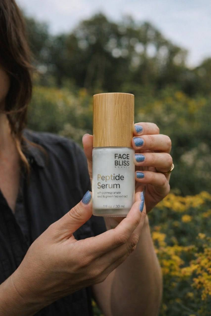 Person holding a 'Face Bliss Peptide Serum' bottle outdoors with greenery in the background